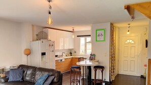 Un salon moderne avec un canapé en cuir, un coin repas avec des tabourets, une cuisine avec des meubles blancs et une grande fenêtre.