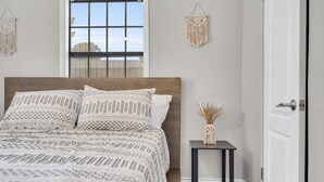 A bedroom with a bed, macrame wall hangings, a small table with a vase, and a window with a view.