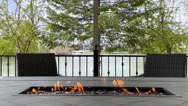 A fire pit with flames, surrounded by stone tiles, overlooking a body of water with a railing and trees in the background. - Lakefront Getaway with Game Room and Theater Seating (Moses Lake)