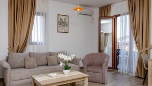 Un salón con un sofá color beige, un sillón rosa, una mesa de centro de madera con un jarrón de flores blancas y un cuadro en la pared.