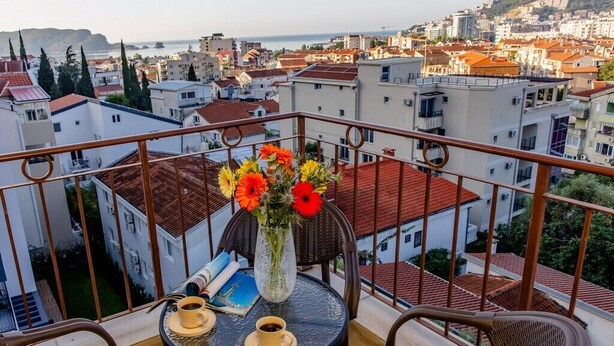 Un balcón con una mesa, sillas, un florero con flores y vistas a un paisaje urbano.