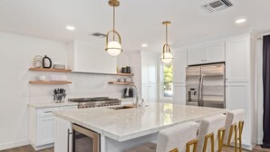 Una cocina moderna con armarios blancos, una isla central con taburetes de bar, electrodomésticos de acero inoxidable e iluminación colgante.
