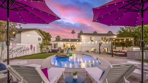 Zona de piscina exterior con sillas reclinables y sombrillas, rodeada de edificios con guirnaldas de luces.