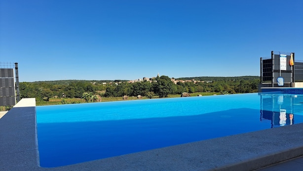 Una piscina rectangular con agua azul cristalina, rodeada por una solana de hormigón. Al fondo, se aprecia un paisaje verde exuberante con árboles y un pueblo lejano.
