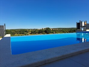 Uma piscina retangular com água azul cristalina, rodeada por um deck de concreto. Ao fundo, vê-se uma paisagem verde exuberante com árvores e uma aldeia distante.
