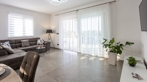 Una sala moderna con un sofá gris, una mesita de centro, una lámpara de pie y un ventanal con cortinas translúcidas.