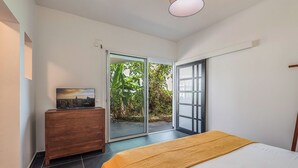 A bedroom with a bed, a wooden dresser, a cityscape painting, and a sliding glass door leading to a garden.