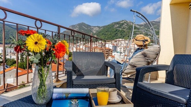 Een balkon met rieten stoelen, een tafel met dienblad, een vaas met bloemen en een stadsgezicht op de achtergrond.