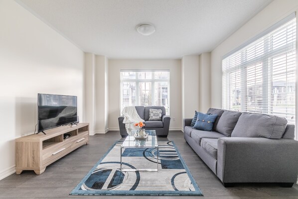 A modern living room with a flat-screen TV, a wooden entertainment unit, a grey sofa, and a patterned rug.