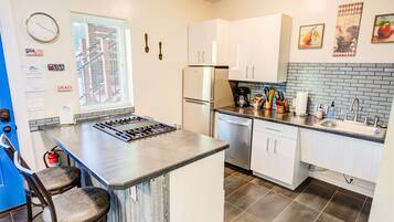 A kitchen with white cabinets, a stainless steel refrigerator, a gas stove, and a sink. There are two bar stools and a window with blinds.