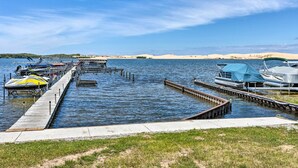 A marina with boats docked at wooden piers, a grassy area in the foreground, and a distant shoreline with sand dunes. - Breezy Silver Lake Getaway: Fishing Dock, Grill! (Mears)