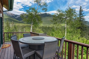 Terraza de madera con una mesa redonda y sillas de mimbre, con vistas a una cadena montañosa boscosa.