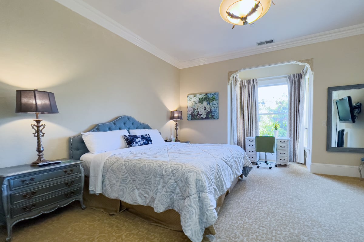 A bedroom with a bed, bedside tables, a lamp, a painting, a window with curtains, and a mirror on the wall.