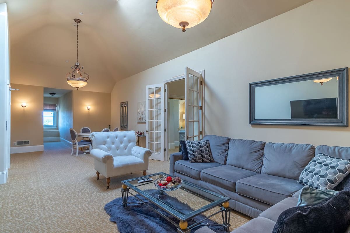 A spacious living room with a grey sofa, a white armchair, and a glass coffee table. There is a large mirror on the wall and a chandelier hanging from the ceiling.