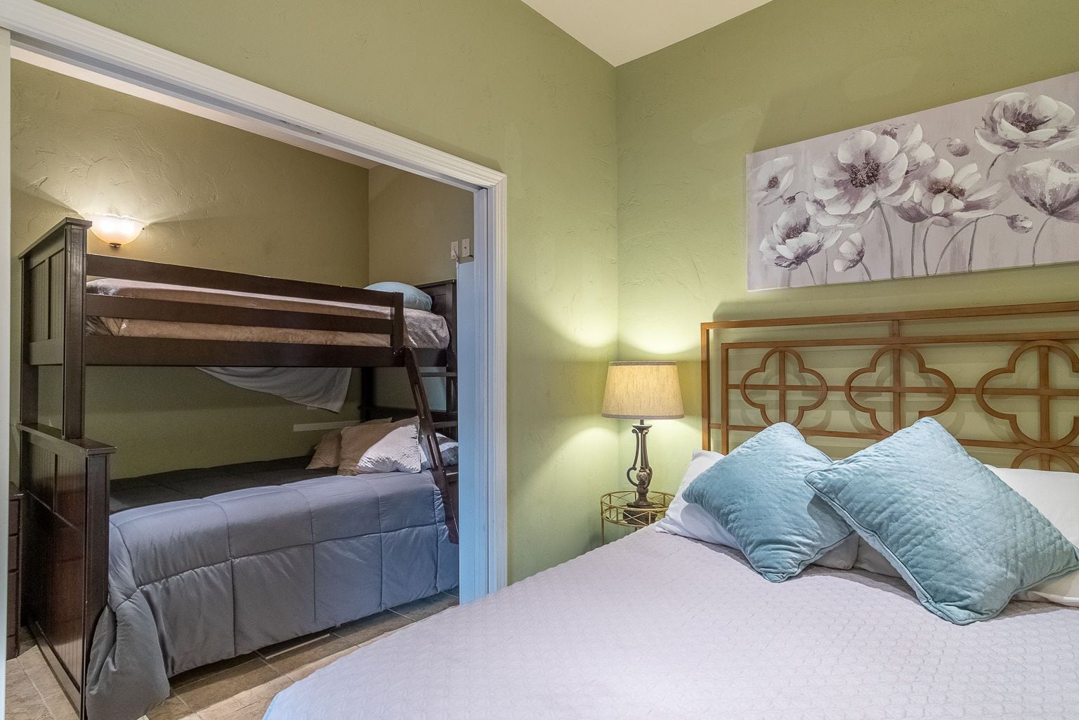 A room with bunk beds, a single bed, a nightstand with a lamp, and a floral painting on the wall.
