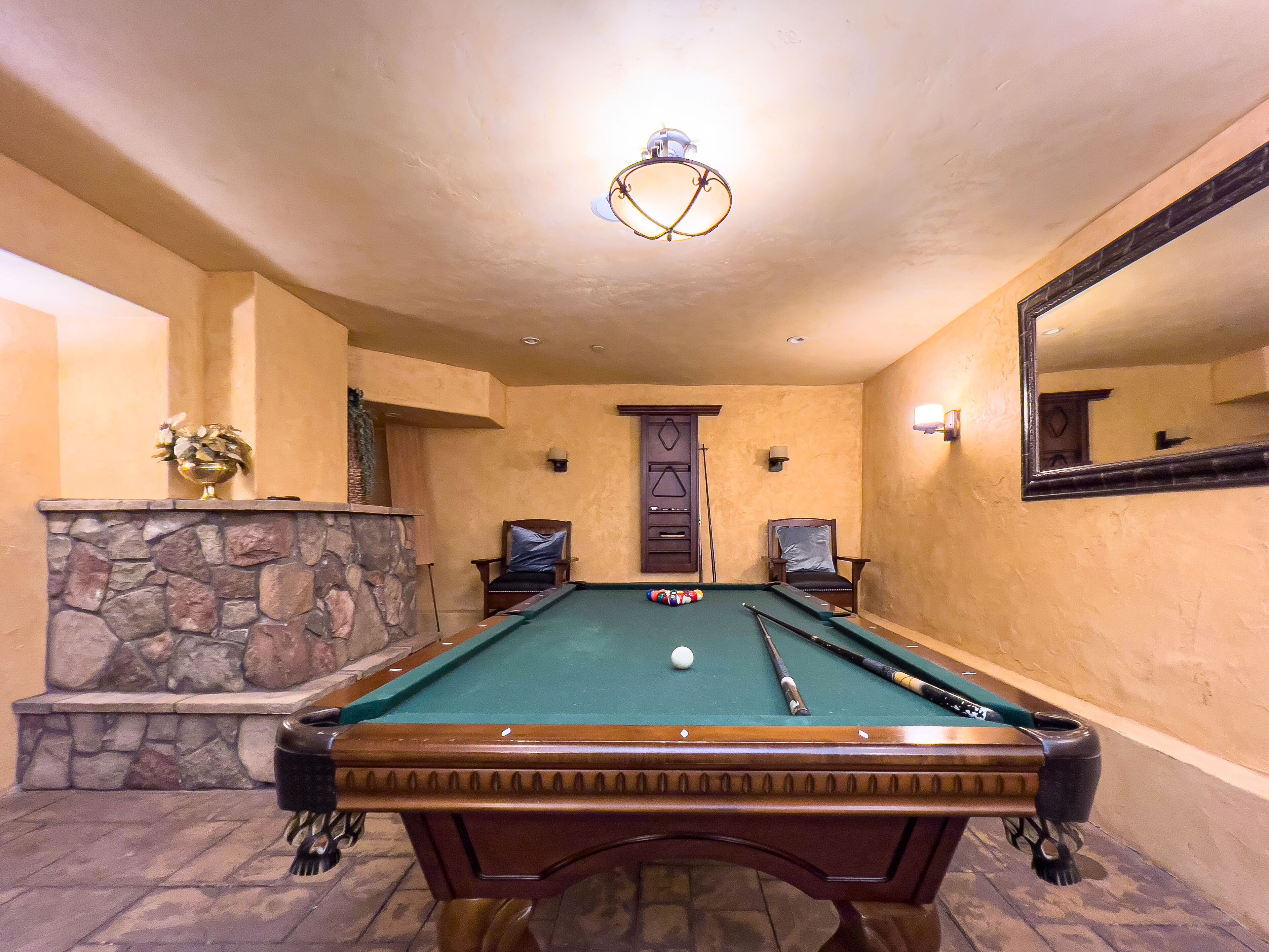 A pool table with a stone fireplace, two armchairs, and a mirror on the wall.