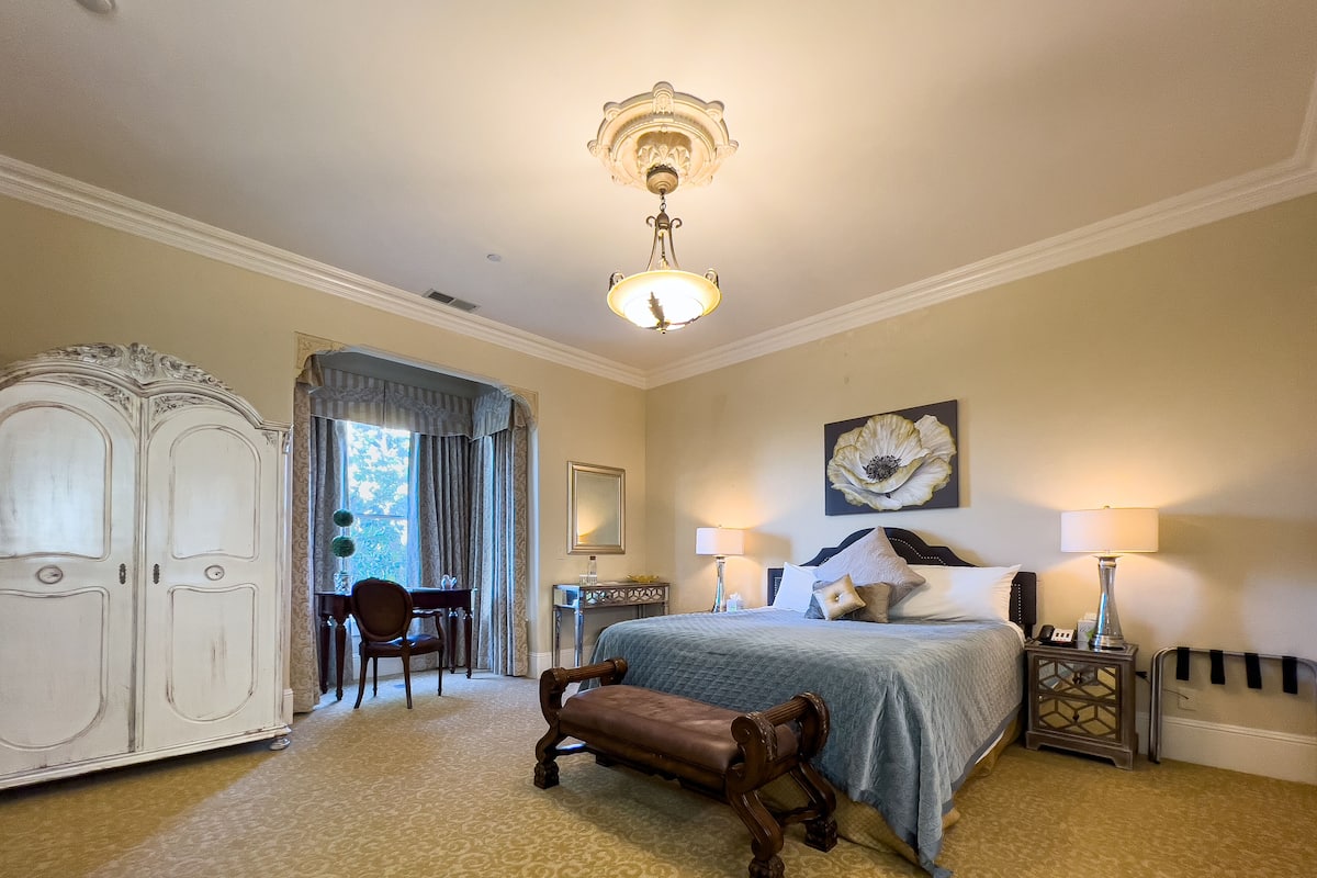 A bedroom with a bed, a nightstand, a lamp, a chair, a wardrobe, and a large window.