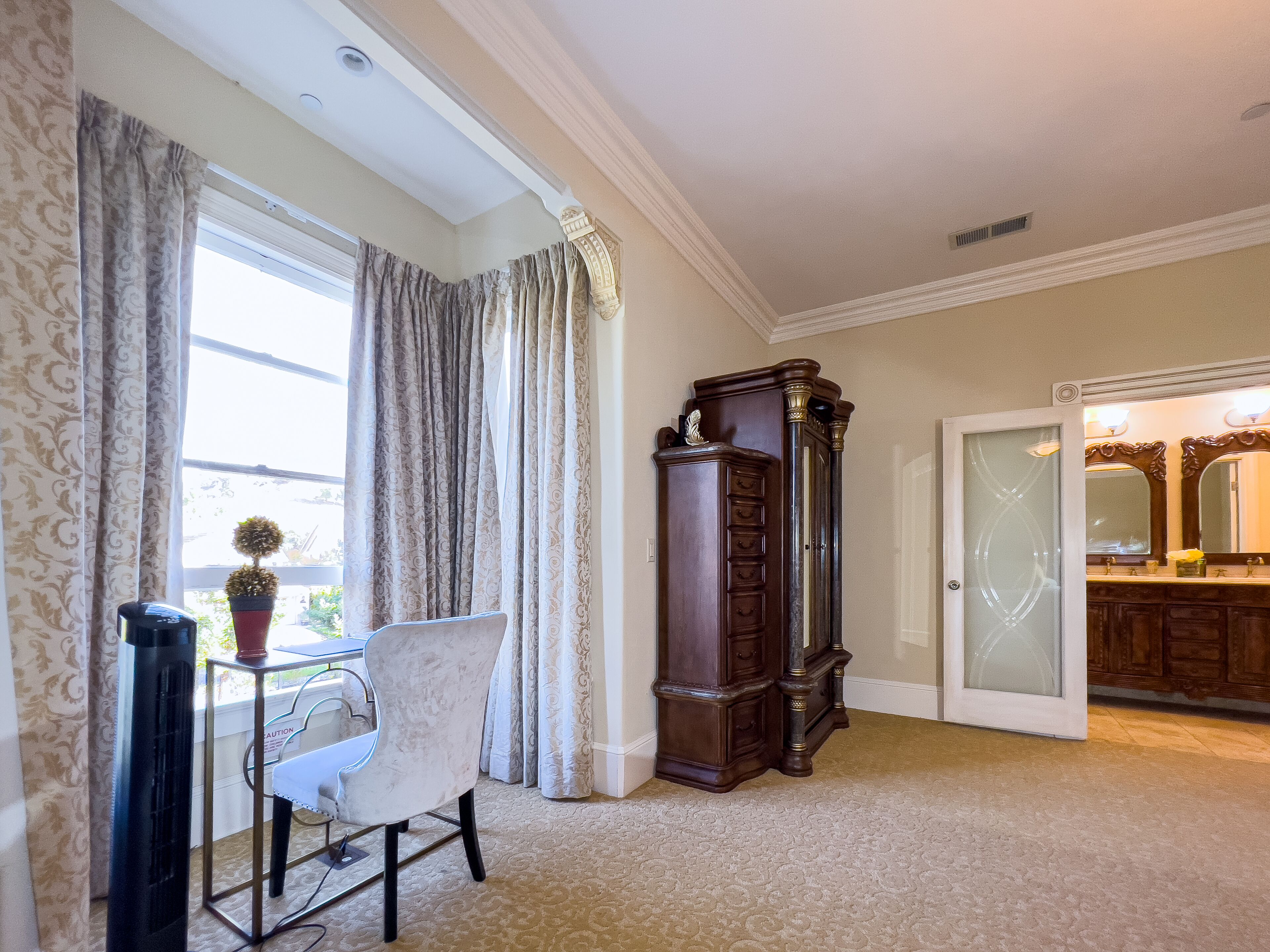 A room with a large window, a tall wooden cabinet, a white chair, and a bathroom with a mirror and sink.