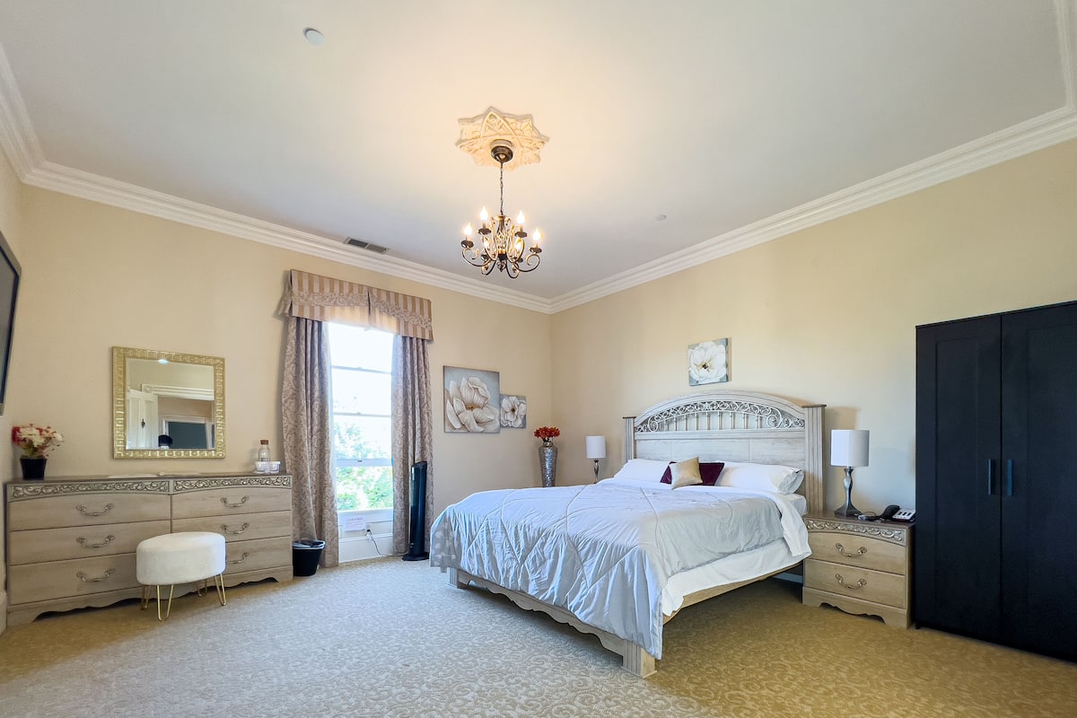A bedroom with a large bed, a dresser, a nightstand, a wardrobe, a chandelier, and a window with curtains.