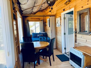 A wooden cabin interior with a dining table, chairs, a sofa with 'BEACH' pillows, a small kitchen area, and a door. - Ocean-view Cabin on the Anchor Point Bluff (Anchor Point)