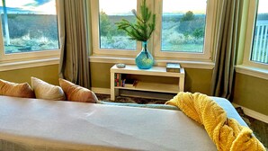 Een slaapkamer met een groot bed, een nachtkastje met een vaas en uitzicht op het landschap door de ramen.