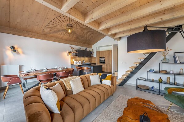 A modern living room with a wooden ceiling, a dining area with a wooden table and chairs, and a staircase with a black railing.
