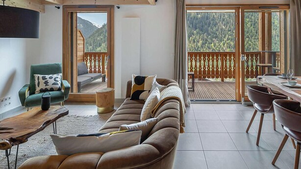 En moderne stue med en sofa, et spisebord dækket til seks personer og en balkon med udsigt over bjergene.