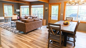 A spacious living area with a sofa, dining table, and chairs, large windows, and a chandelier. - Luxury Lakefront Retreat | Sauna, Dock & Kayaks (Talkeetna)