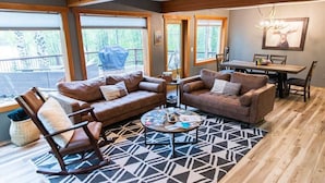 A cozy living room with a sectional sofa, a rocking chair, and a coffee table. There is a dining table with chairs, a chandelier, and a large window with a view of the outdoors. - Luxury Lakefront Retreat | Sauna, Dock & Kayaks (Talkeetna)