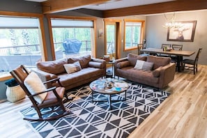 A cozy living room with a sectional sofa, a rocking chair, and a coffee table. There is a dining table with chairs, a chandelier, and a large window with a view of the outdoors.