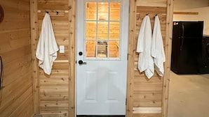A wooden cabin with a white door, two towels hanging on hooks, and a black refrigerator to the right. - Luxury Lakefront Retreat | Sauna, Dock & Kayaks (Talkeetna)