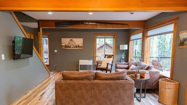 A cozy living room with a brown sofa, a TV, and a window overlooking a cabin. - Luxury Lakefront Retreat | Sauna, Dock & Kayaks (Talkeetna)