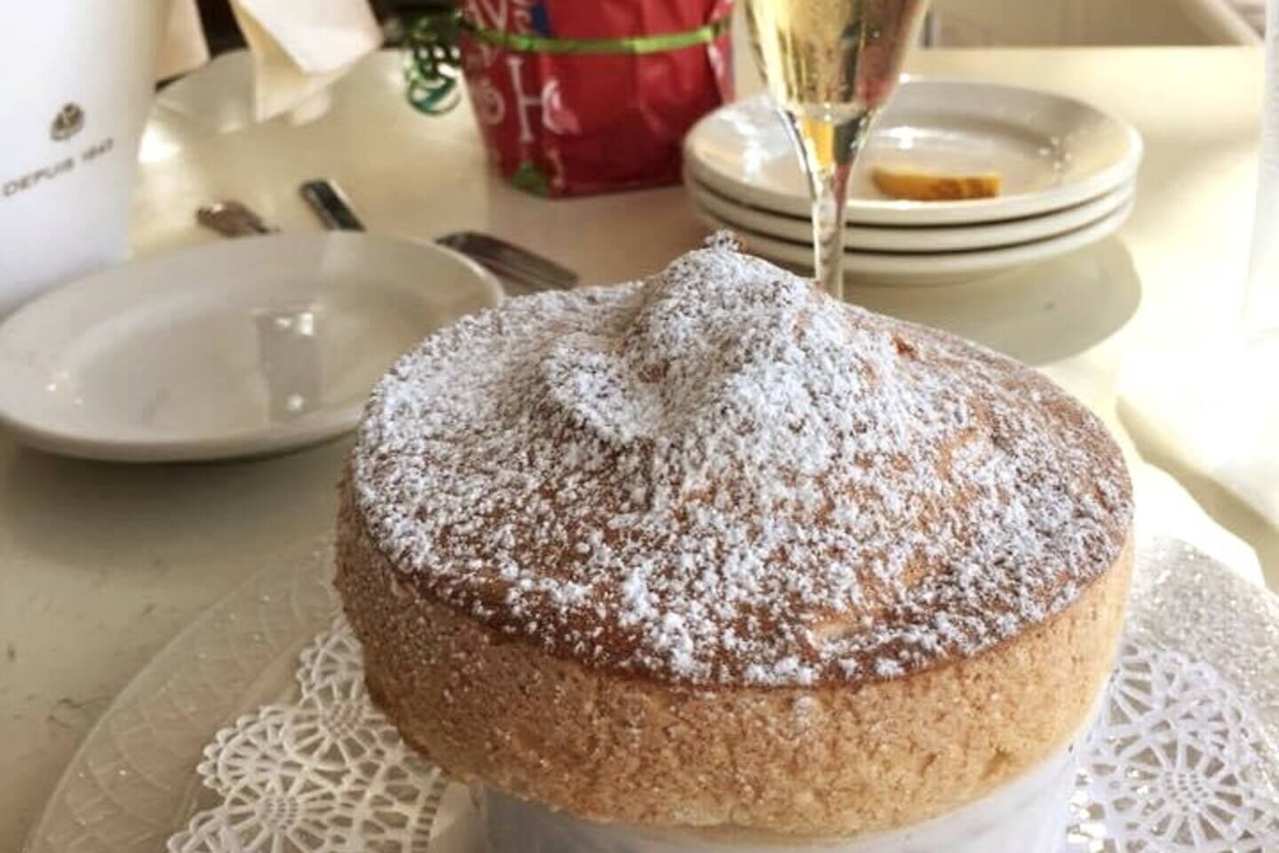 A round cake with powdered sugar on a white plate, a glass of champagne, and a red gift box in the background.