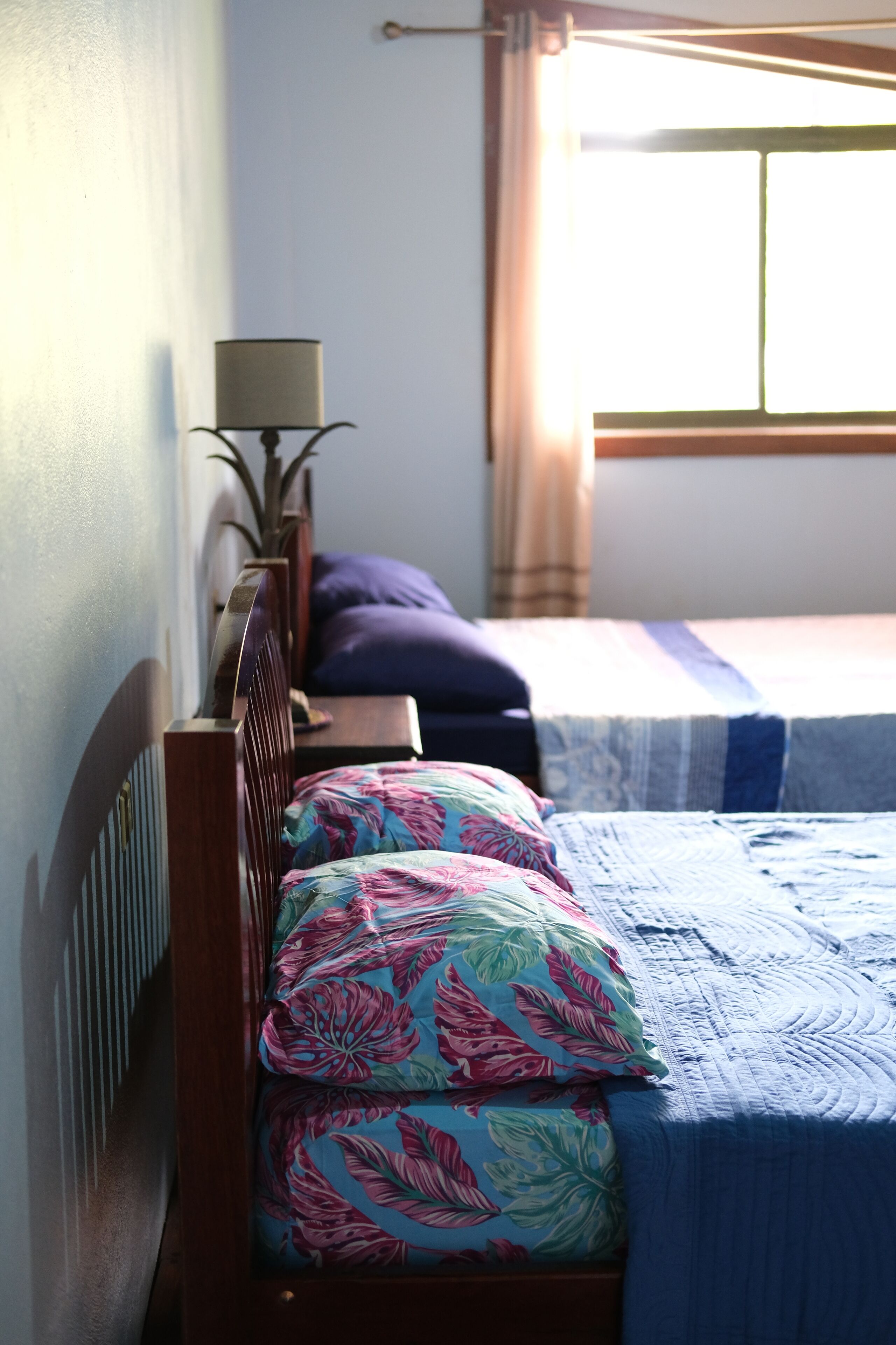 Una cama con un cojín estampado con motivos florales, una colcha azul y una lámpara de noche.