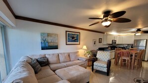 A living room with a beige sectional sofa, a dining area with wooden chairs, a ceiling fan, and a kitchen with white cabinets and appliances.