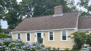 Une maison à deux étages avec une cheminée en briques, un porche à l’avant et un jardin bien entretenu agrémenté de buissons d’hortensias.