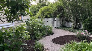Un chemin de jardin recouvert de gravier, bordé de différentes plantes et entouré d’une clôture en bois peinte en blanc.