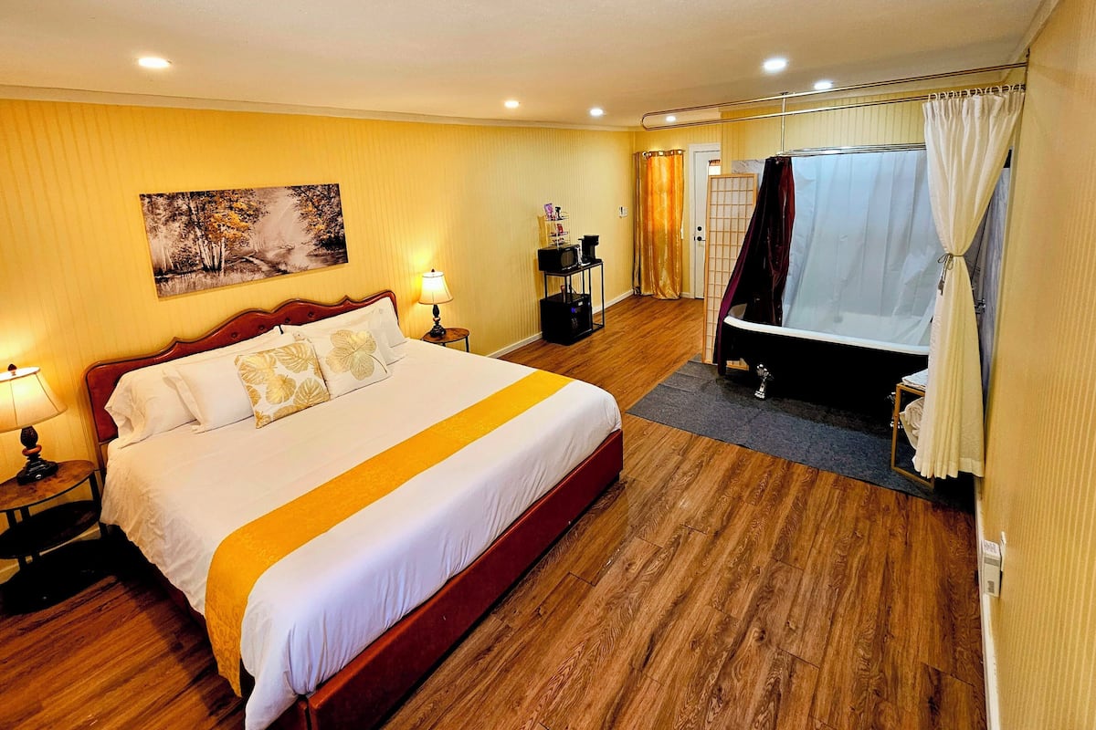 A hotel room with a large bed, a bedside table with a lamp, a small table with a TV, a bathtub, and a shower curtain.