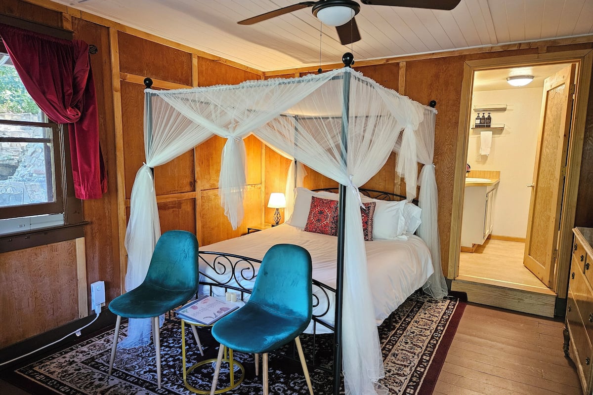 A bedroom with a four-poster canopy bed, two blue chairs, a small table, and a patterned rug.