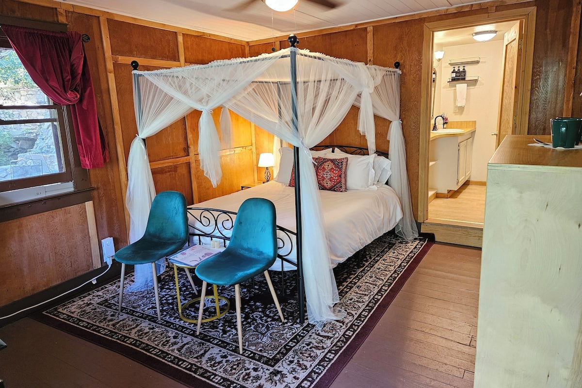 A room with a four-poster bed, two blue chairs, a small table, a patterned rug, wooden walls, a ceiling fan, and a bathroom visible through an open door.