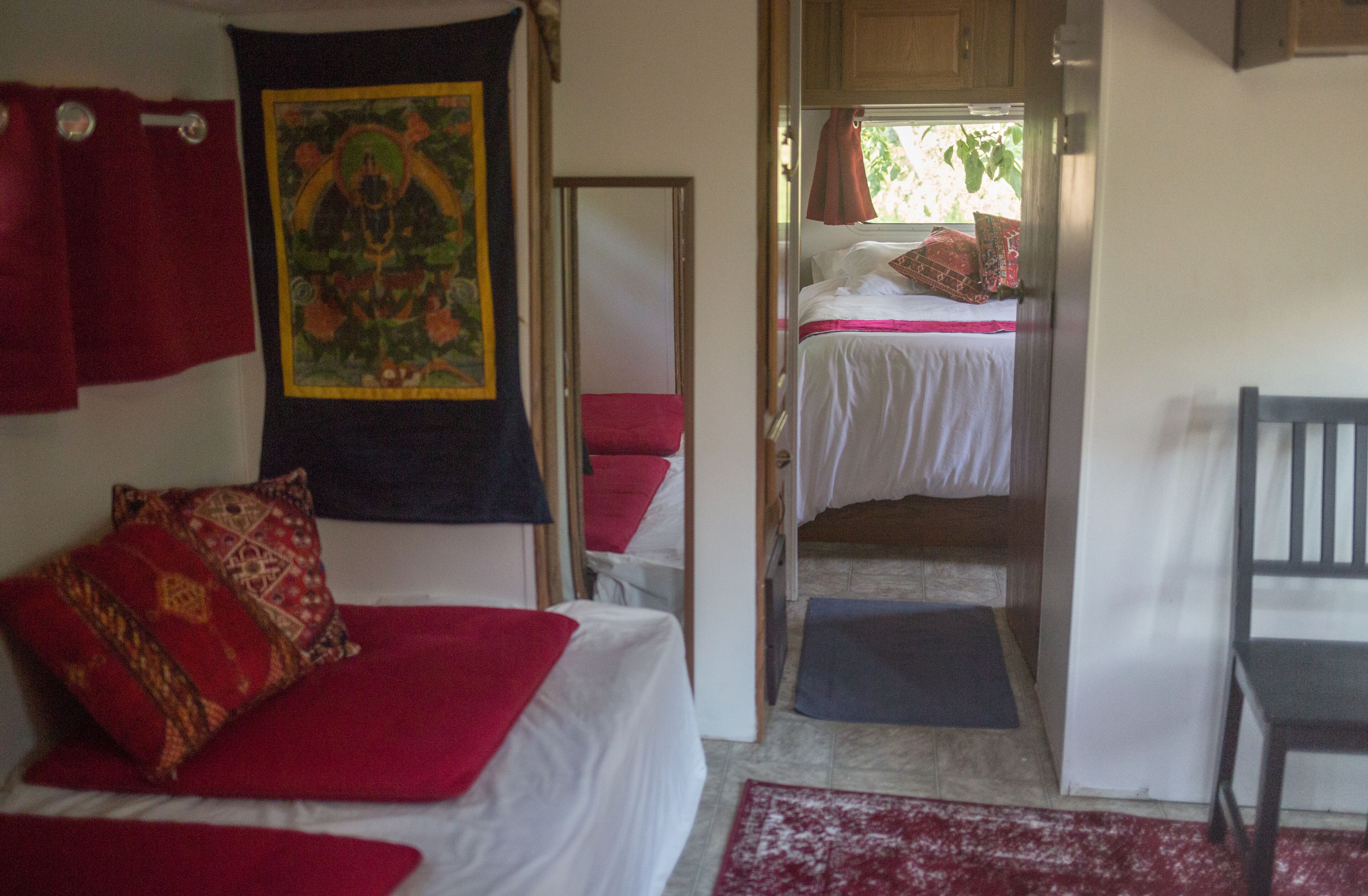 Una habitación con dos camas, una con ropa de cama roja y la otra con ropa de cama blanca. Hay una cortina roja, un tapiz y una silla de madera.