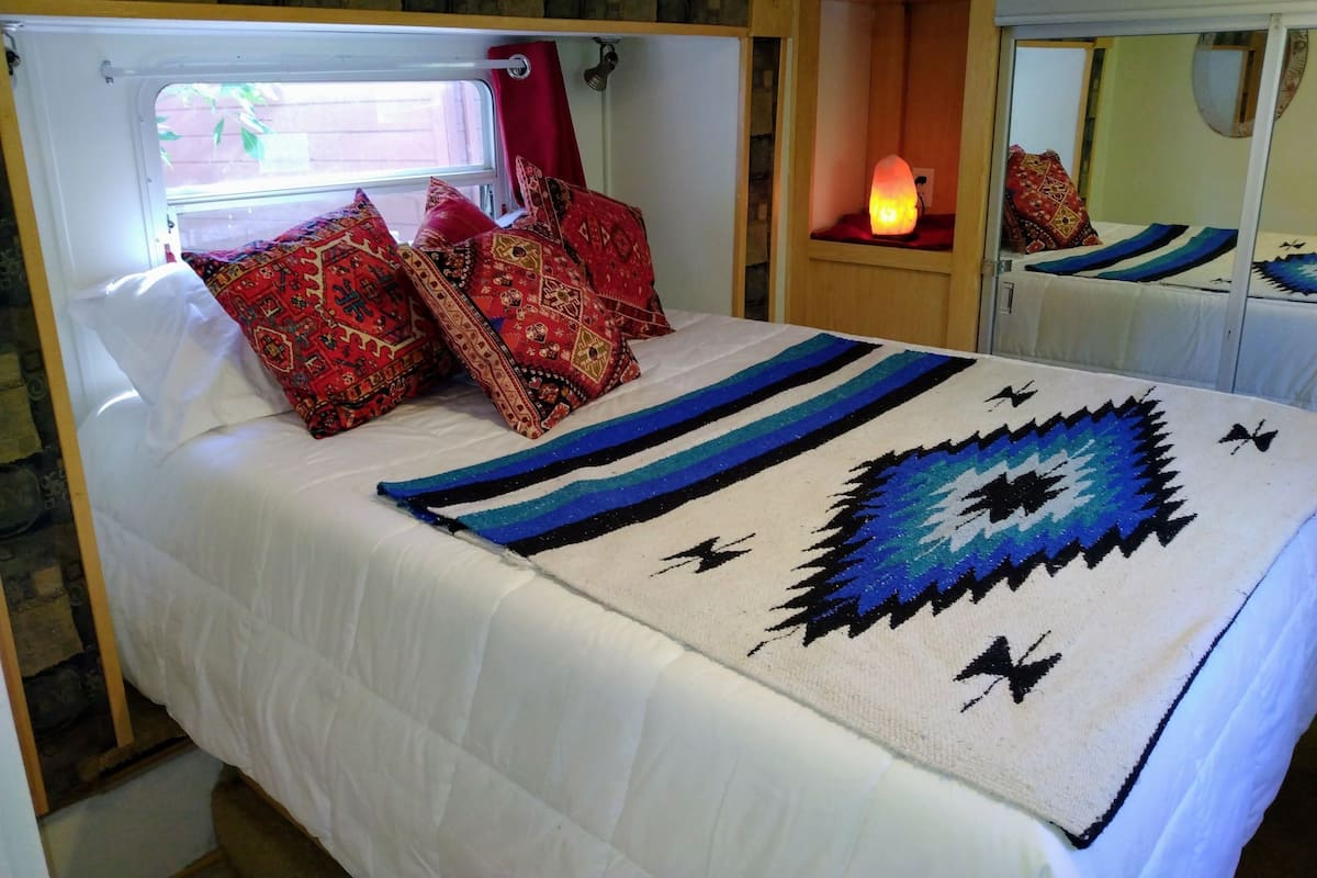 A bed with a white comforter, red and patterned pillows, a blue and black blanket, and a small bedside lamp.