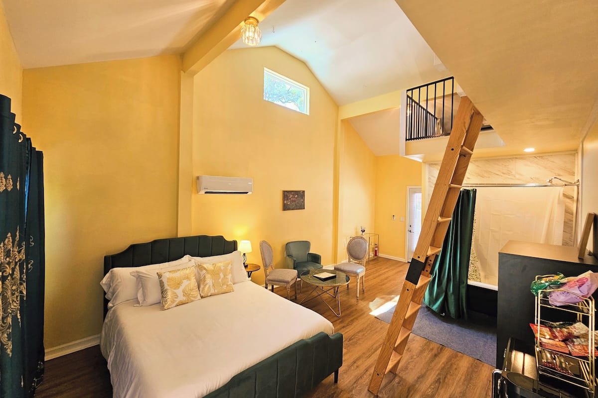 A bedroom with a bed, a chair, a small table, a desk, a staircase, and an air conditioning unit.