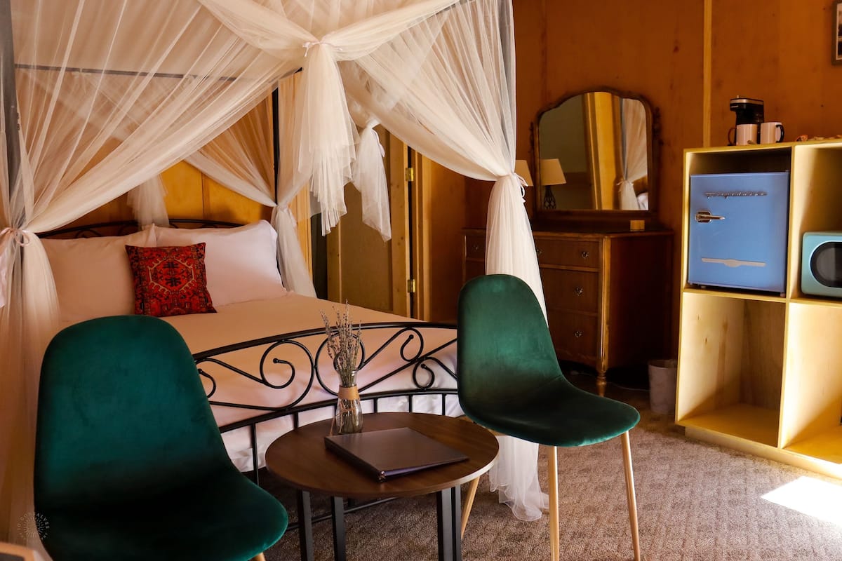 A bedroom with a canopy bed, two green chairs, a small table with a book and vase, a dresser, and a blue cabinet.