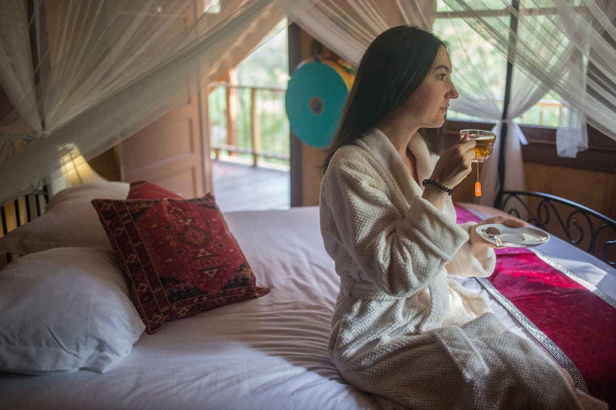 A person sitting on a bed with a red and white patterned pillow, holding a glass of drink.