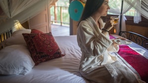 Una persona sentada en una cama con un almohadón de estampado rojo y blanco, sosteniendo un vaso de bebida.