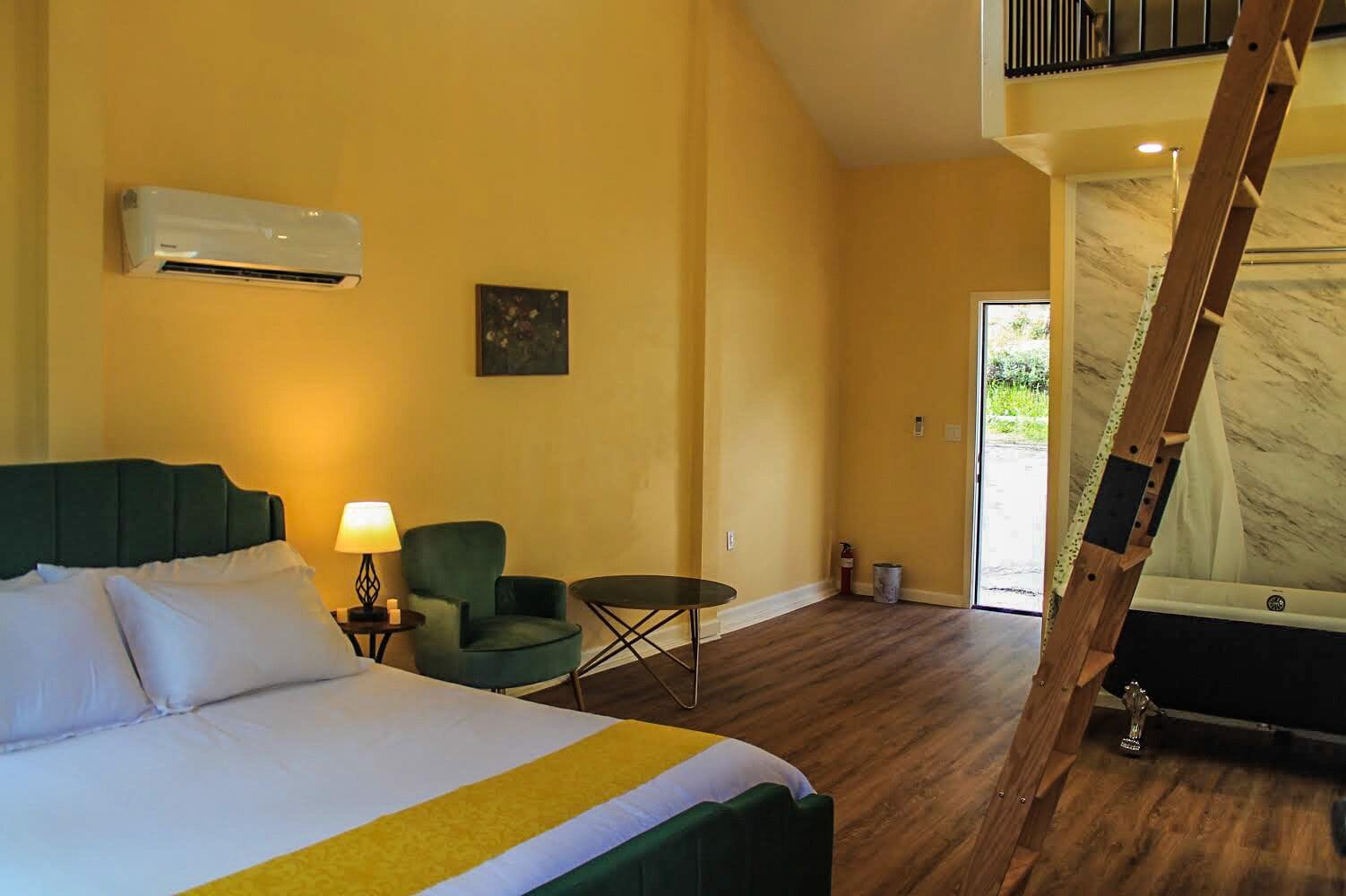 A hotel room with a bed, a green armchair, a small table, and a ladder leading to a balcony.
