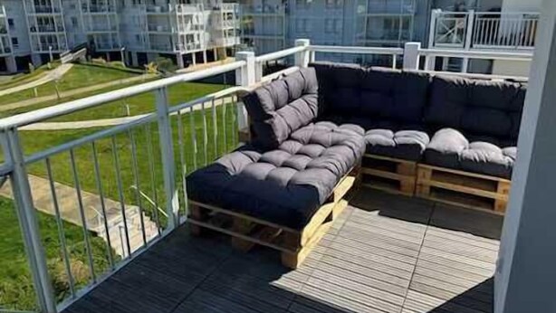 A balcony with a sectional sofa, a wooden deck, and a view of apartment buildings. - Appartement Conquistar - Welkeys (Cabourg)