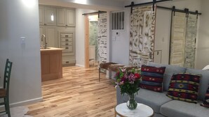 Ein Wohnzimmer mit einer Couch, einem kleinen Tisch mit einer Vase und Holzboden. Es gibt eine Küchenzeile mit Schränken und einem Spülbecken.