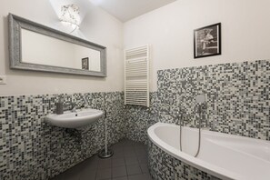 Une salle de bain équipée d’un lavabo, d’un miroir et d’une baignoire, avec des murs recouverts de mosaïques et un radiateur blanc.